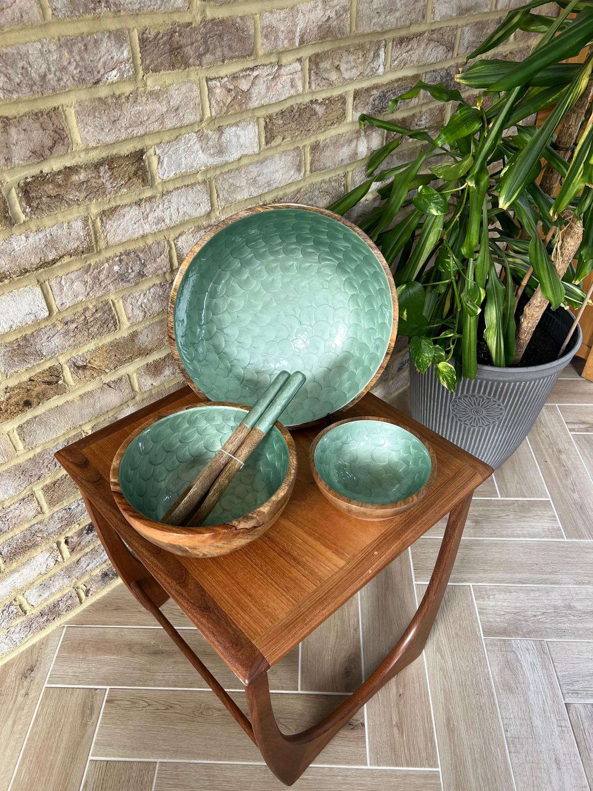 Three hand painted wooden bowls in a light green tea colour, displayed on a wooden side table next to a potted plant against a brick wall. With matching salad servers made from sustainable mango wood.