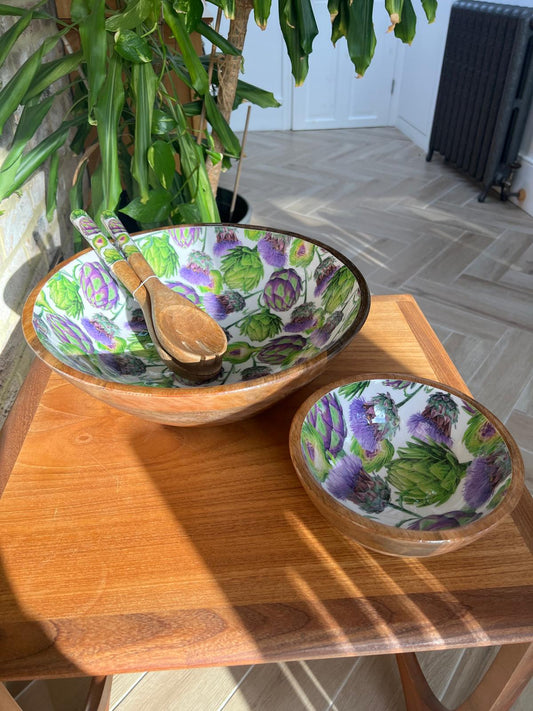 A small and large round wooden salad bowl decorated with a repeating pattern of green artichokes and purple thistles on a white background. A matching wooden serving spoon and fork rest inside the bowl, which is placed on a wooden table against a brick wall background.  Made from sustainable mango wood. Dimensions: Large: Diameter 35cm, depth 15cm
Small: Diameter 18cm, 10cm deep