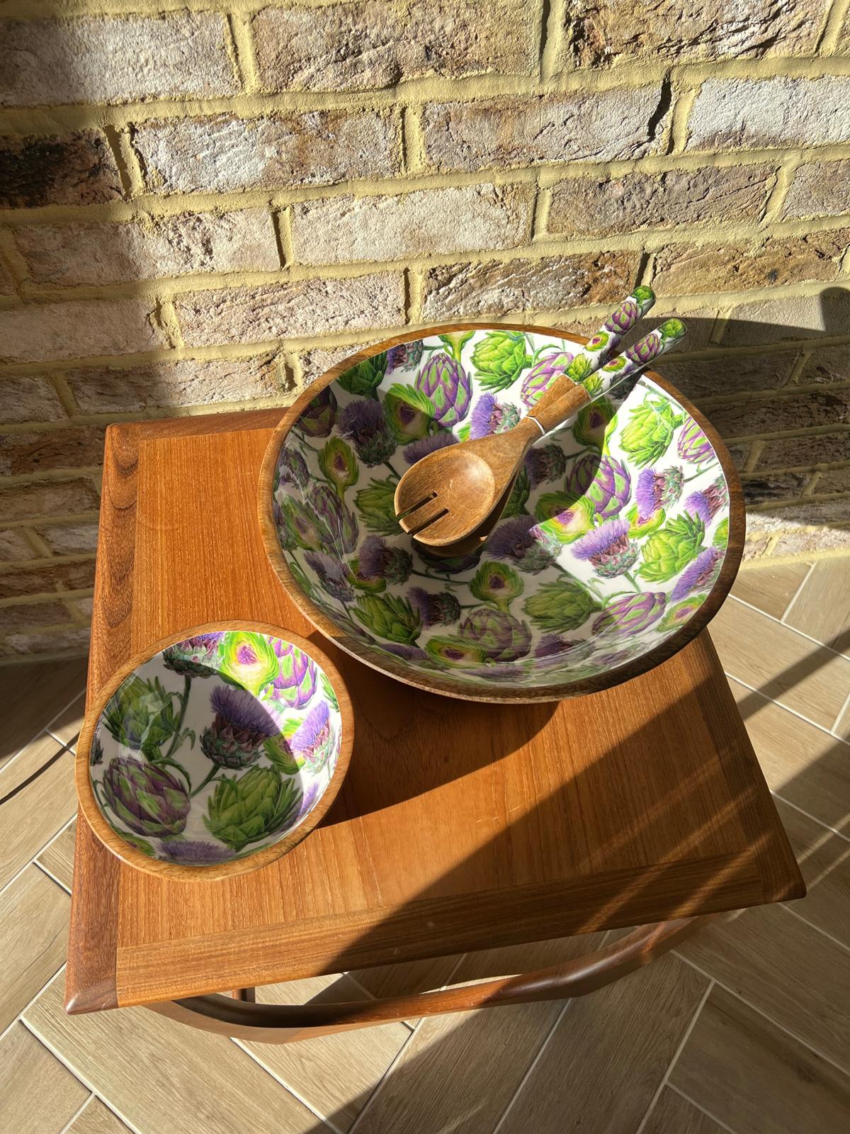 A small and large round wooden salad bowl decorated with a repeating pattern of green artichokes and purple thistles on a white background. A matching wooden serving spoon and fork rest inside the bowl, which is placed on a wooden table against a brick wall background.  Made from sustainable mango wood. Dimensions: Large: Diameter 35cm, depth 15cm
Small: Diameter 18cm, 10cm deep