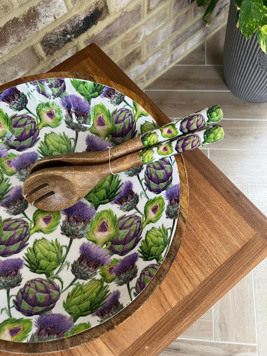 A large wooden salad bowl decorated with a repeating pattern of green & purple artichokes &  thistles. A matching wooden serving spoon & fork rest inside the bowl.  Made from sustainable mango wood. Dimensions: Diameter 35cm, depth 15cm