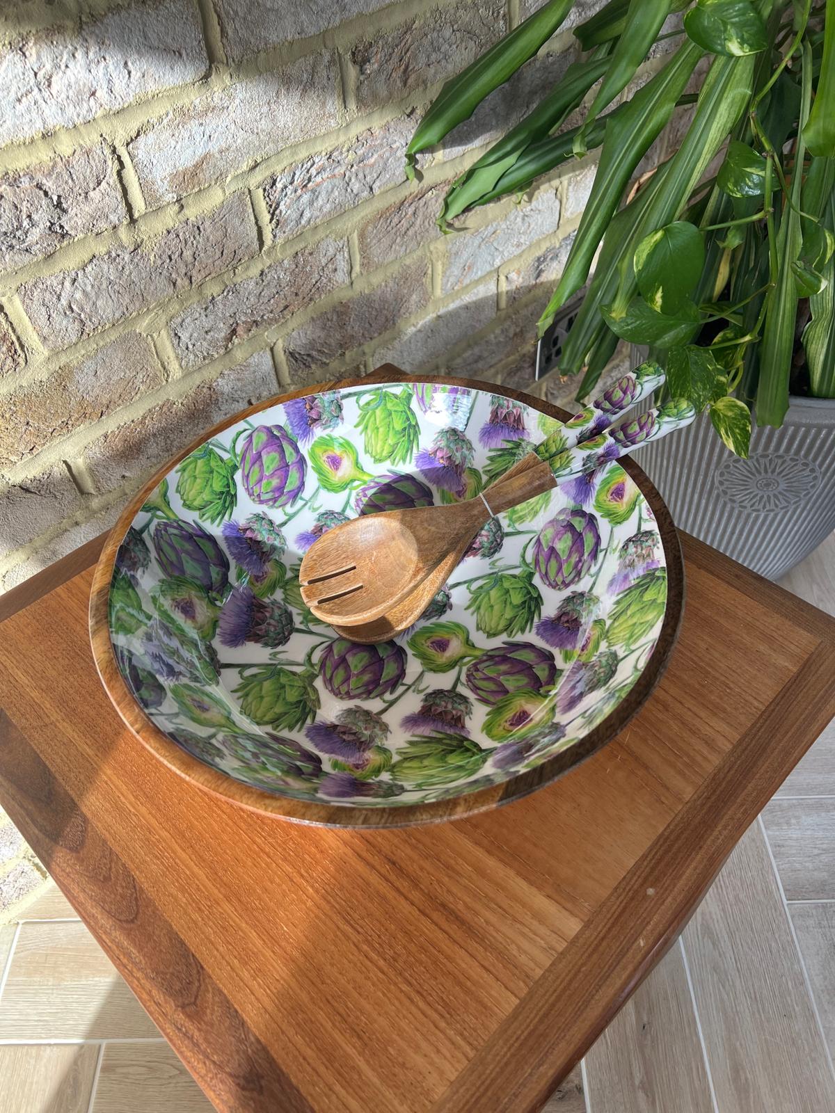 A large round wooden salad bowl decorated with a repeating pattern of green artichokes and purple thistles on a white background. A matching wooden serving spoon and fork rest inside the bowl, which is placed on a wooden table against a brick wall background.  Made from sustainable mango wood. Dimensions: Diameter 35cm, depth 15cm