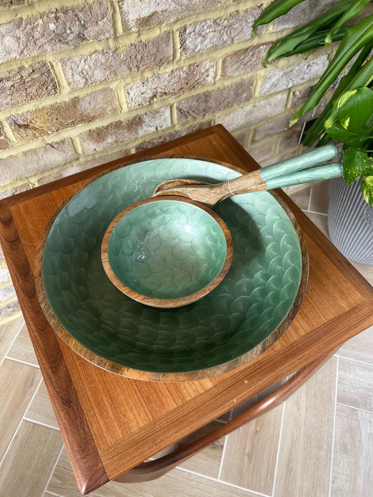 Large and small bowls with a hand painted green tea coloured interior and matching salad servers, set on a wooden table against a brick wall with a green plant in the background.  Made from sustainable mango wood.  Suitable as a unique chip and dip set