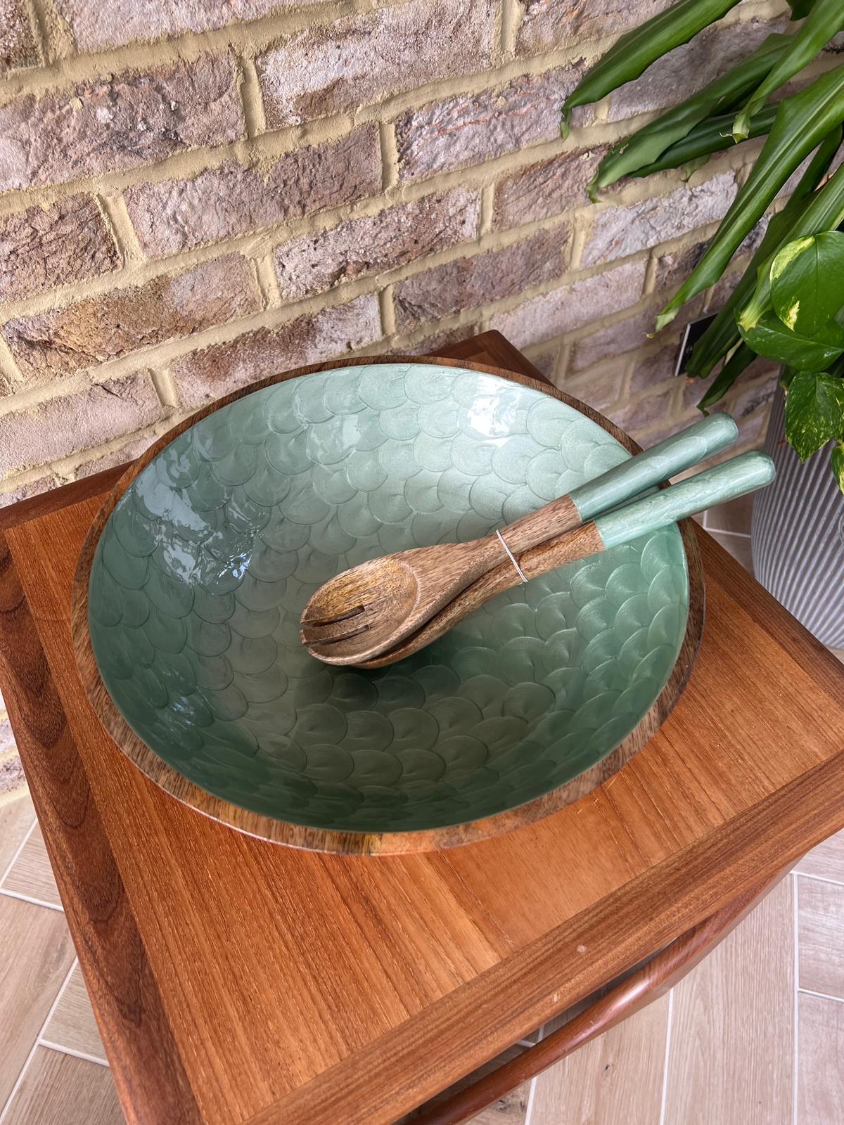 A large wooden salad bowl set with matching salad servers, set on a wooden table against a brick wall with a green plant in the background.  Hand painted and handmade from sustainable mango wood.  Large salad bowl set.  Diameter 38cm, depth 15cm