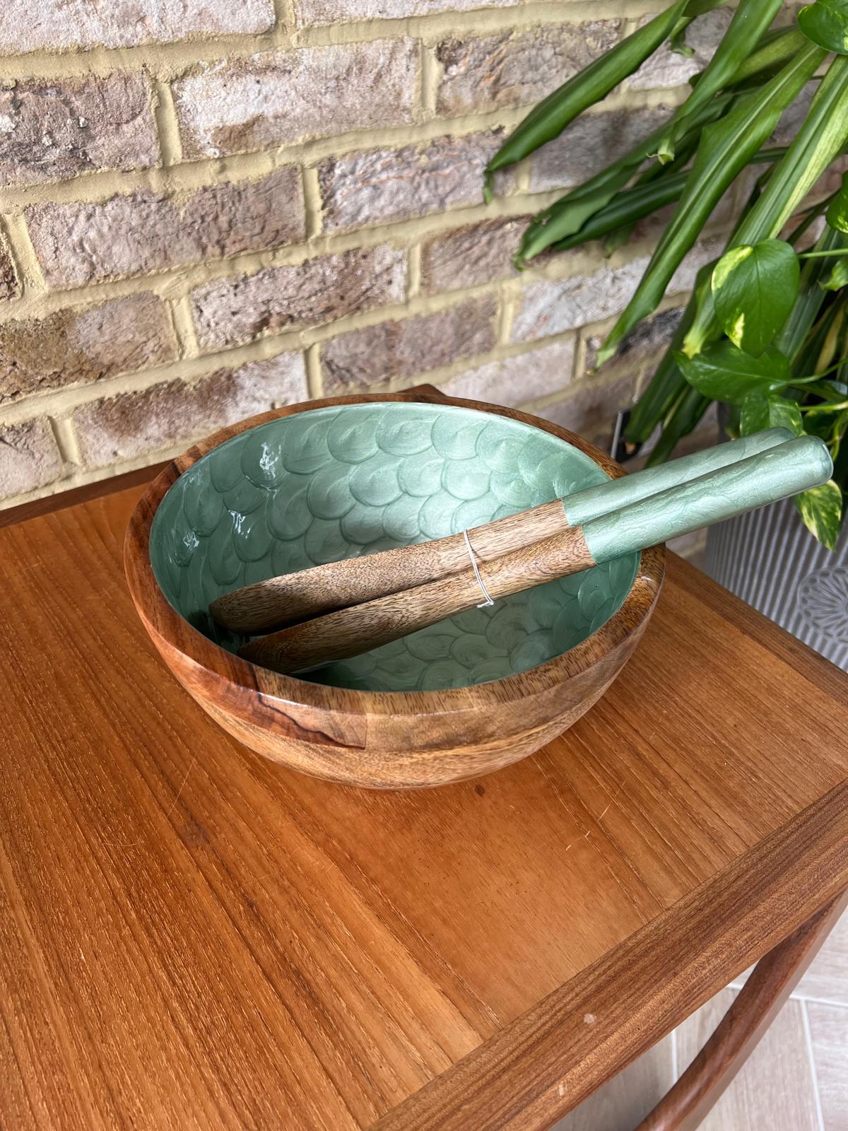 A wooden bowl with a hand painted green tea coloured interior with matching salad servers, set on a wooden table against a brick wall with a green plant in the background.  Made from sustainable mango wood.  Mdeium salad bowl set.  Diameter 25cm, depth 15cm