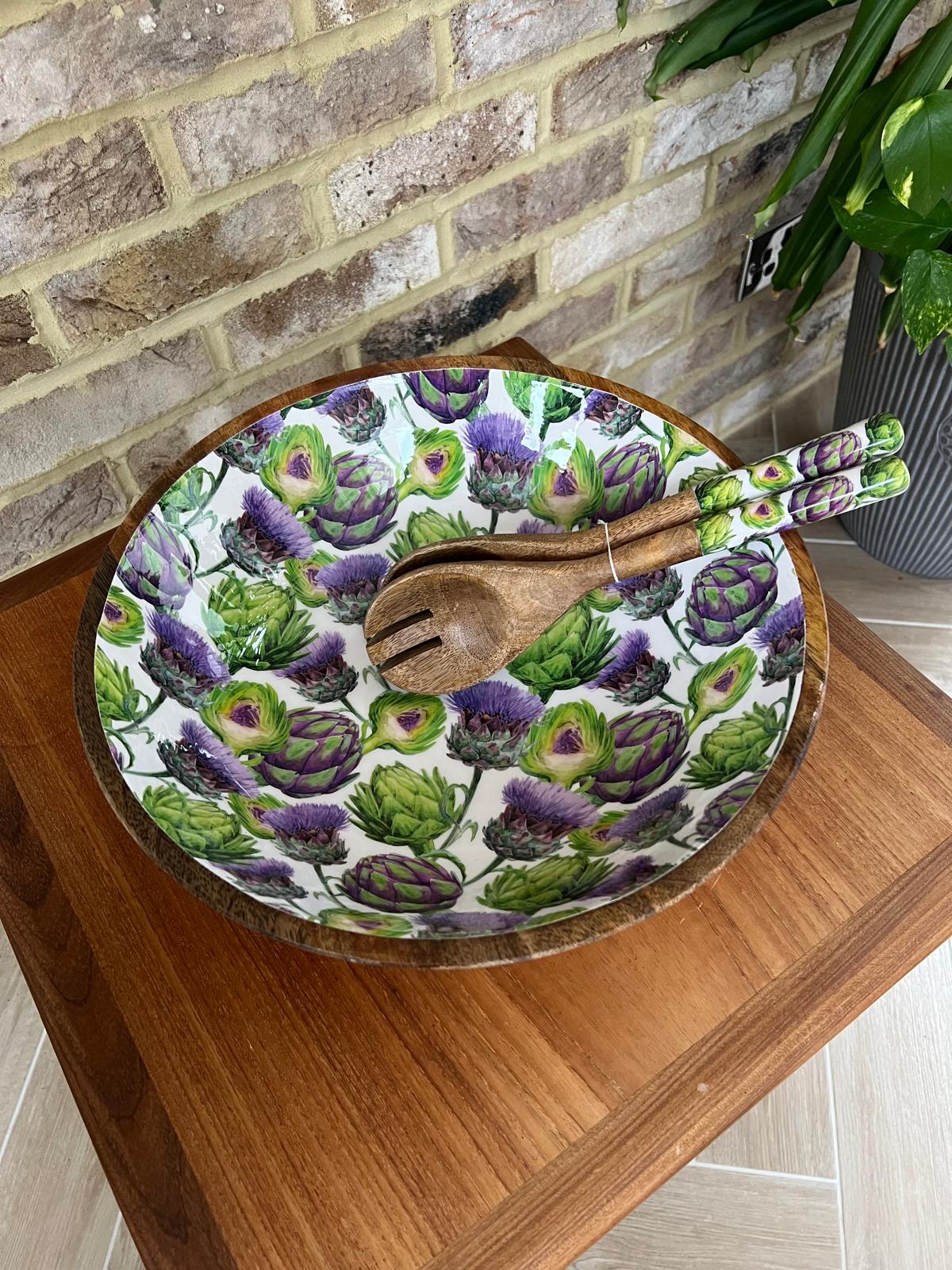 A large round wooden salad bowl decorated with a repeating pattern of green artichokes and purple thistles on a white background. A matching wooden serving spoon and fork rest inside the bowl, which is placed on a wooden table against a brick wall background.  Made from sustainable mango wood. Dimensions: Diameter 35cm, depth 15cm