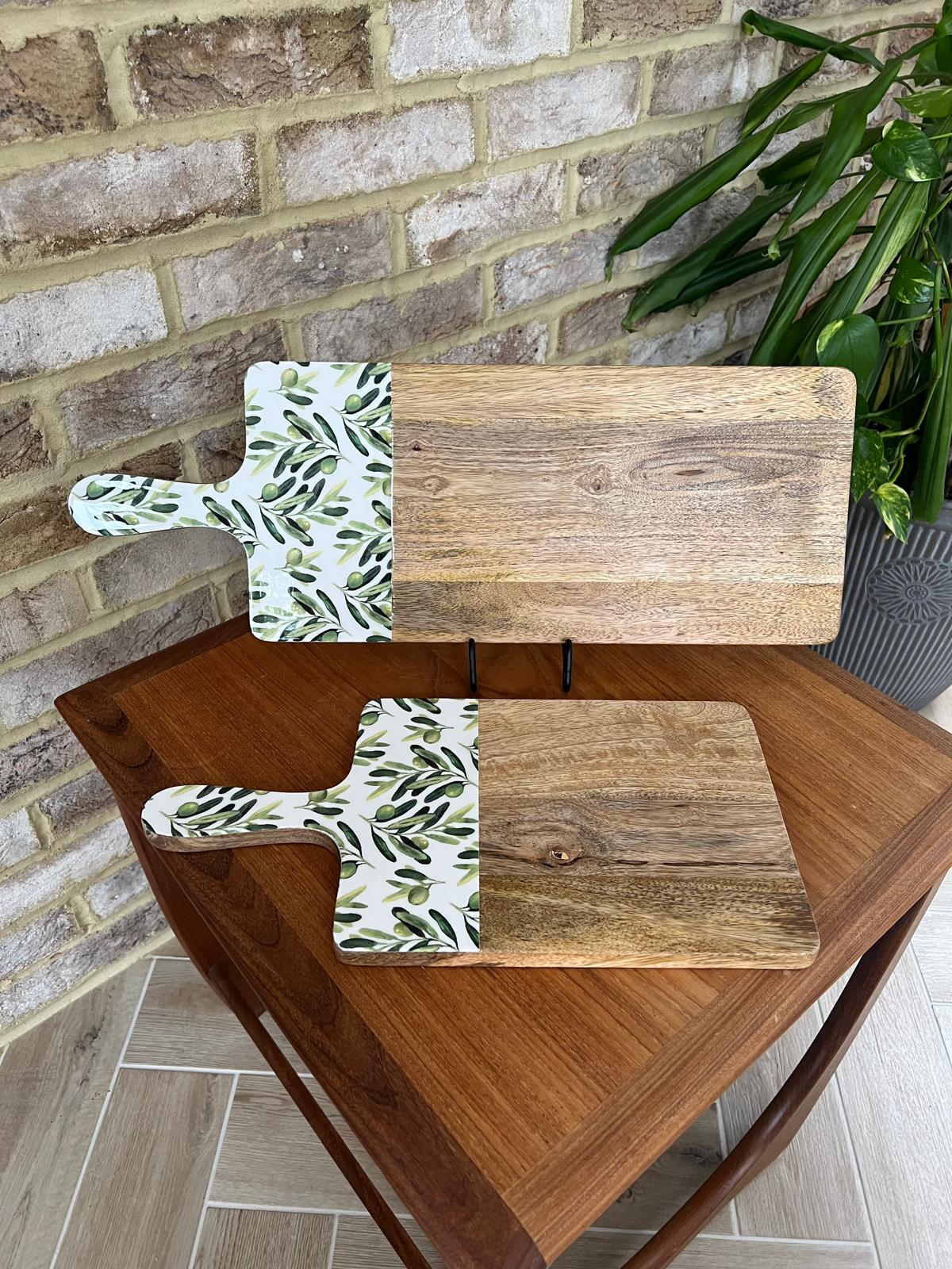 2 wooden serving boards with a white handle featuring a green olive branch pattern, resting on a larger wooden table next to a brick wall and potted plant.  Made from sustainable mango wood.  In medium and large sizes. Suitable for use as a chopping, serving or cheese board.
